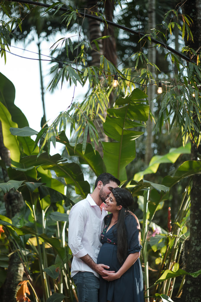 Man and his pregnant wife embrace under banana leaves at the Bamboo Gallery for their maternity session