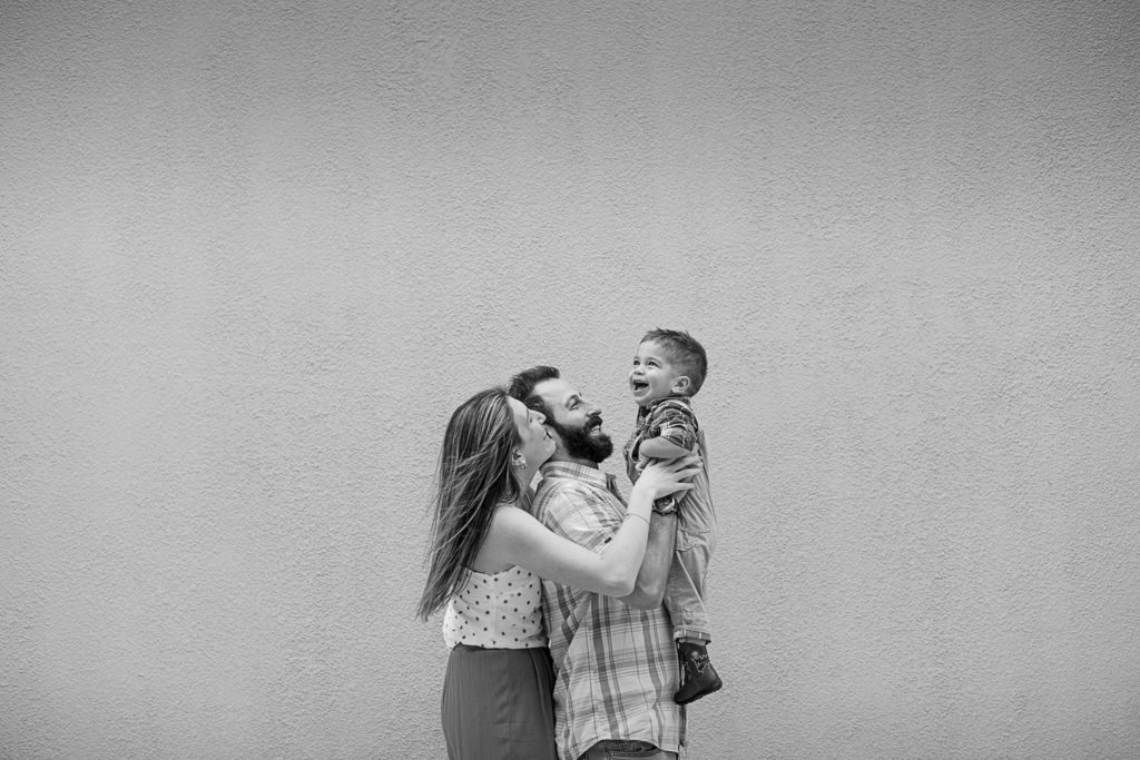 Black and white photo of a mom, dad, and their toddler son against a blank wall at Mizner Park 