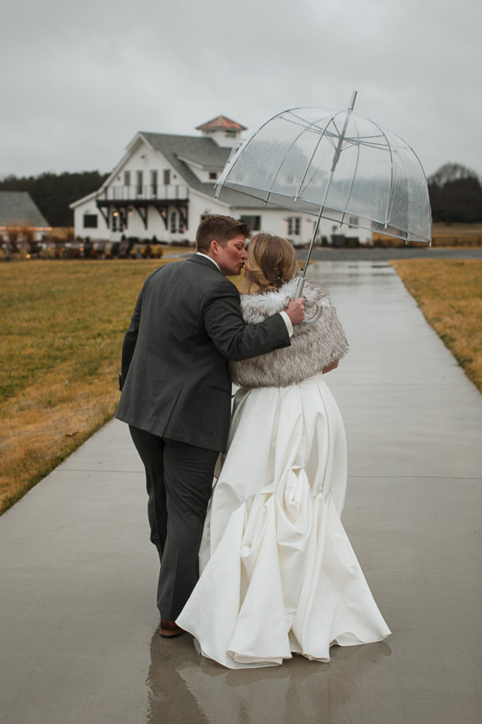 Bride and bride kiss under a clear umbrella on the way to the reception at Chickadee Hill Farms