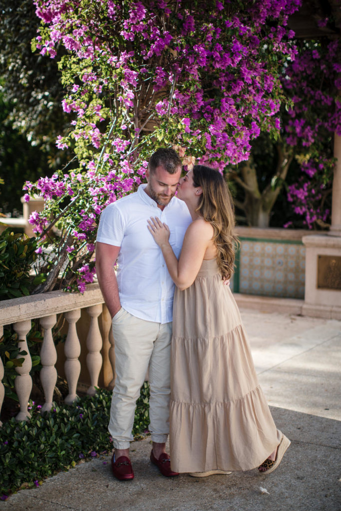 bride-to-be wearing a tan dress kisses the cheek of her future husband in front of a Palm beach courtyard Miami Proposal Photographer