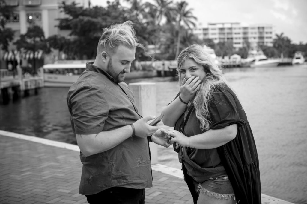 Surprised fiance covers her mouth in disbelief as her partner photographs the ring on his cell phone
