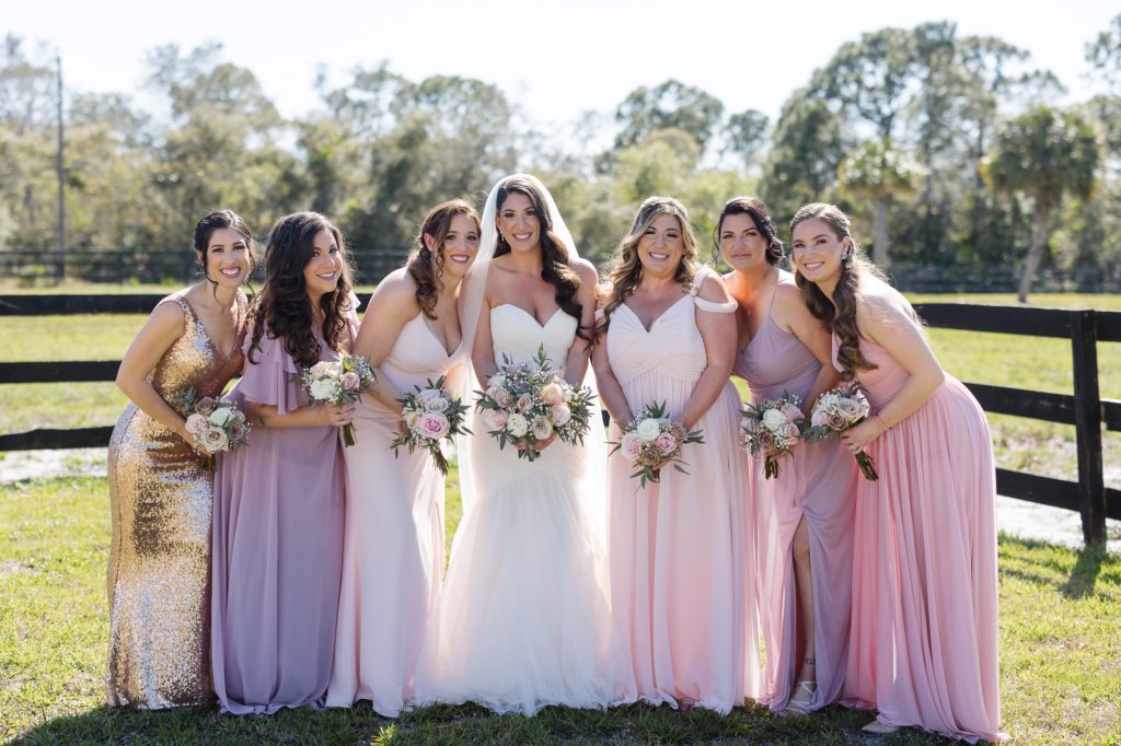 Bridesmaids dressed in pinks, blushes and sequin pose with bride near the fence outside Ever After Farms wedding barn