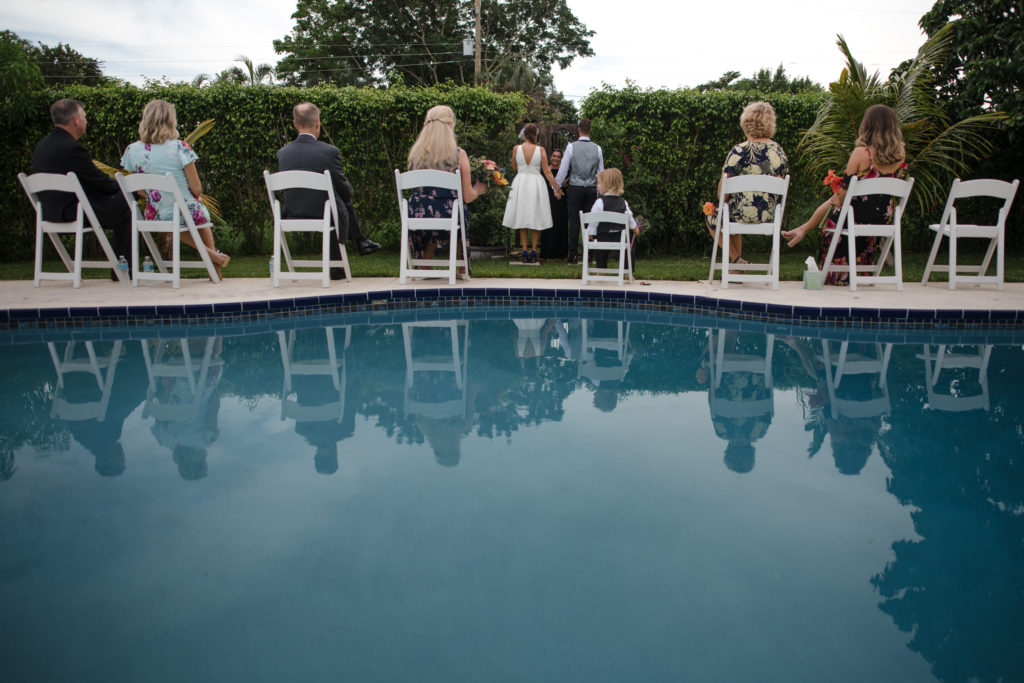 Bride and groom holding hands while their family watches their intimate ceremony with a pool at South Florida Wedding Locations