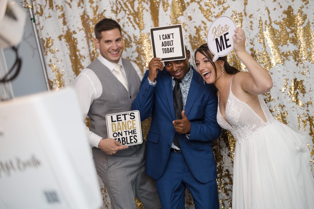 Juju Booth photo booth with a gold and white sequin backdrop. Posing for a photo is the bride and groom with their DJ Glenn Blackk in the center holding fun photo booth prop signs. 