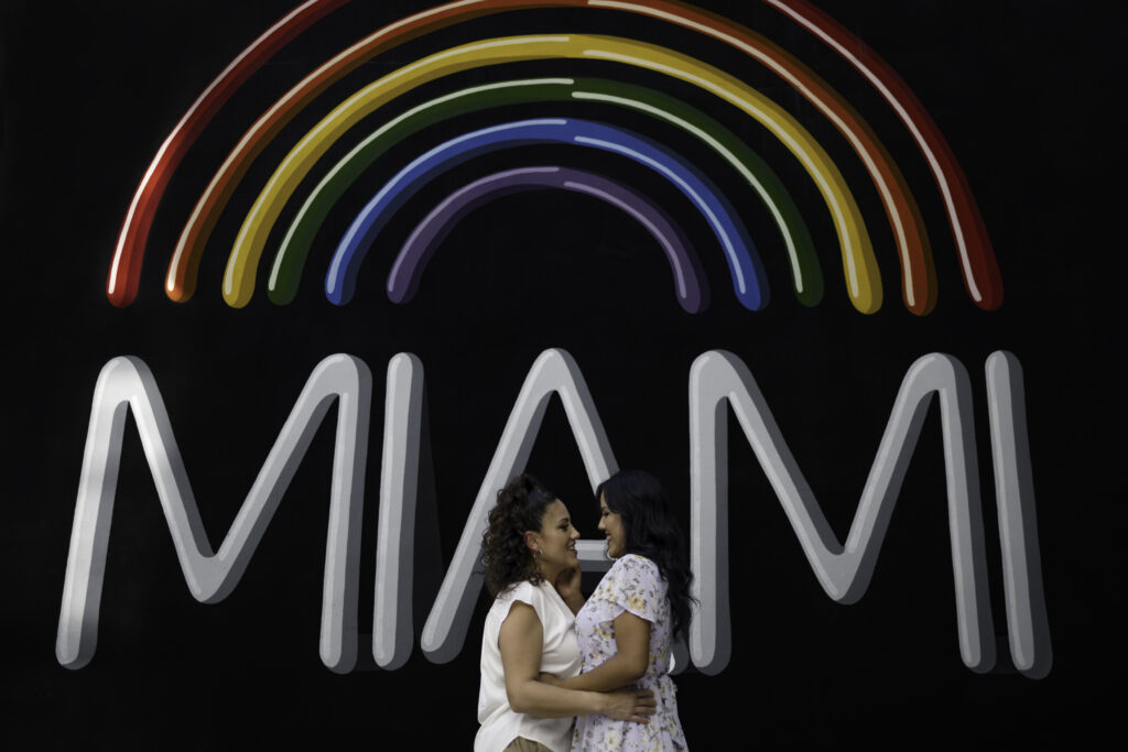 Lesbian couple, both brunettes, pose for an image as they embrace in front of a rainbow mural with the word "MIAMI" under the rainbow