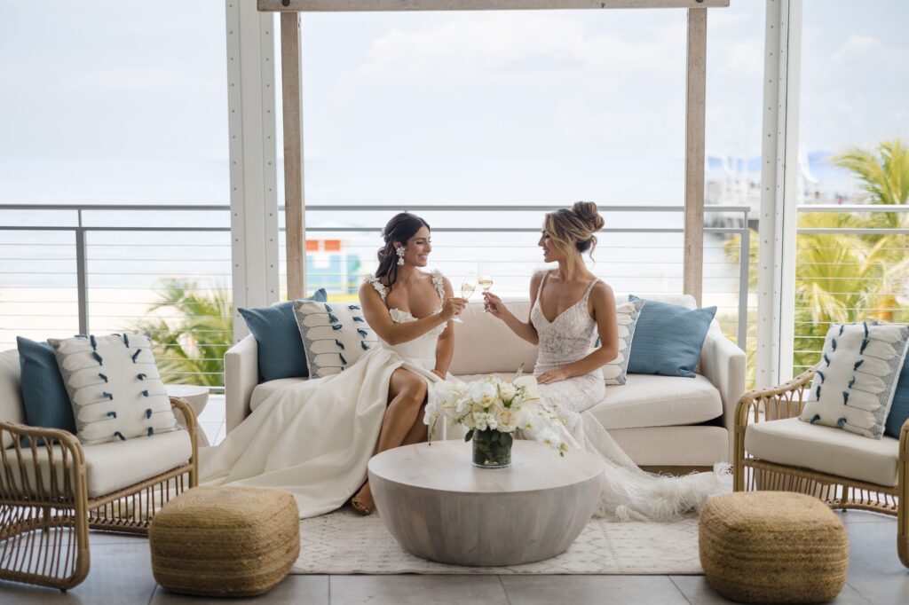 Photo of two brides clicking champagne glasses as they sit on an ivory couch. This couch is part of a lounge area set up for a wedding day and has two additional rattan chairs and ottomans. Furniture is rented from Unearthed Rentals.